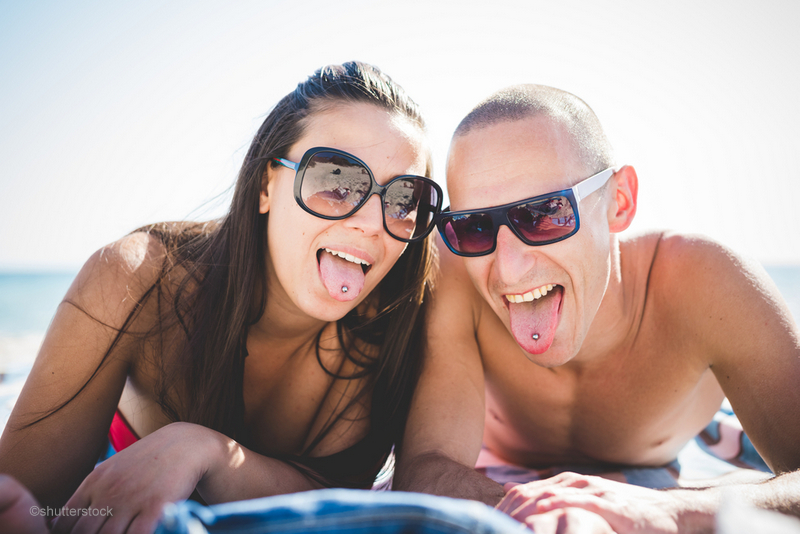 Young couple on the beach sticking out their tongues with oral piercings